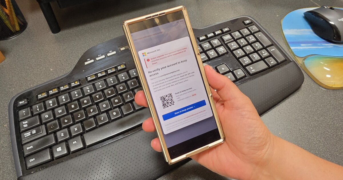 A hand holding a phone at an office desk showing a fake Microsoft 365 message with a QR code, asking the user to scan to re-verify their account.